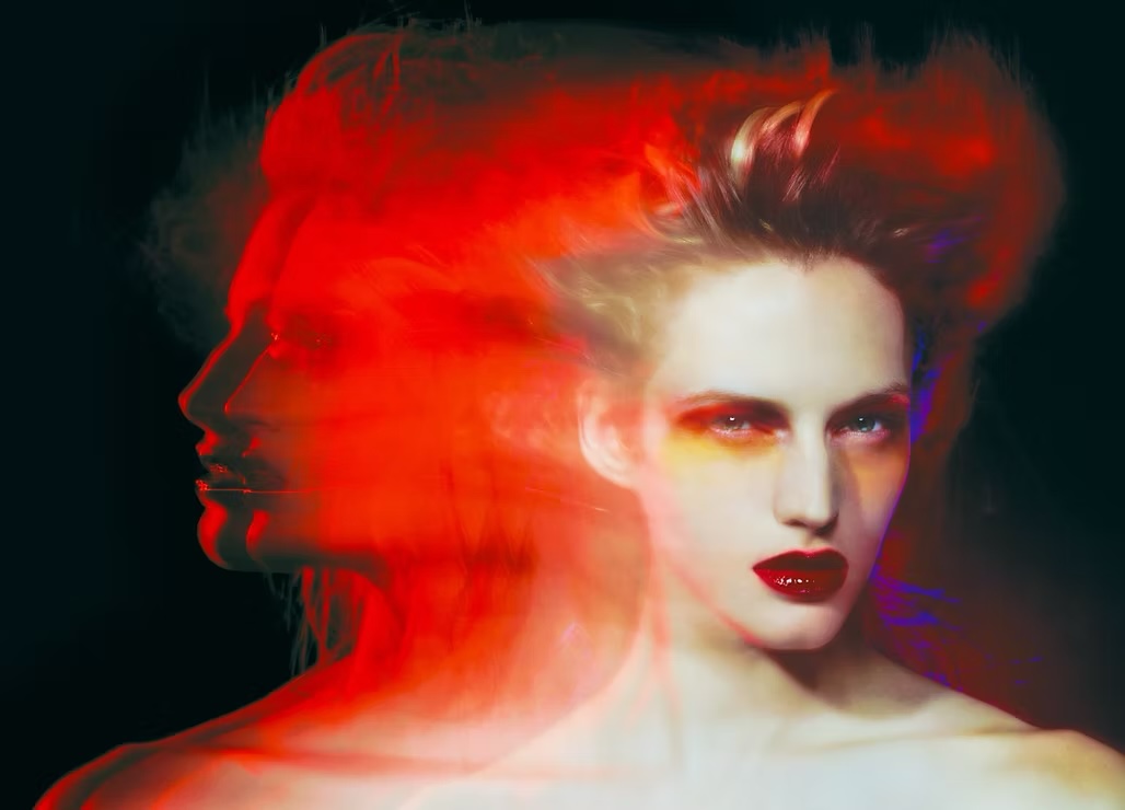 Surreal portrait of a woman with red lips and hair, distorted with a side profile overlay against a dark, blurred background.