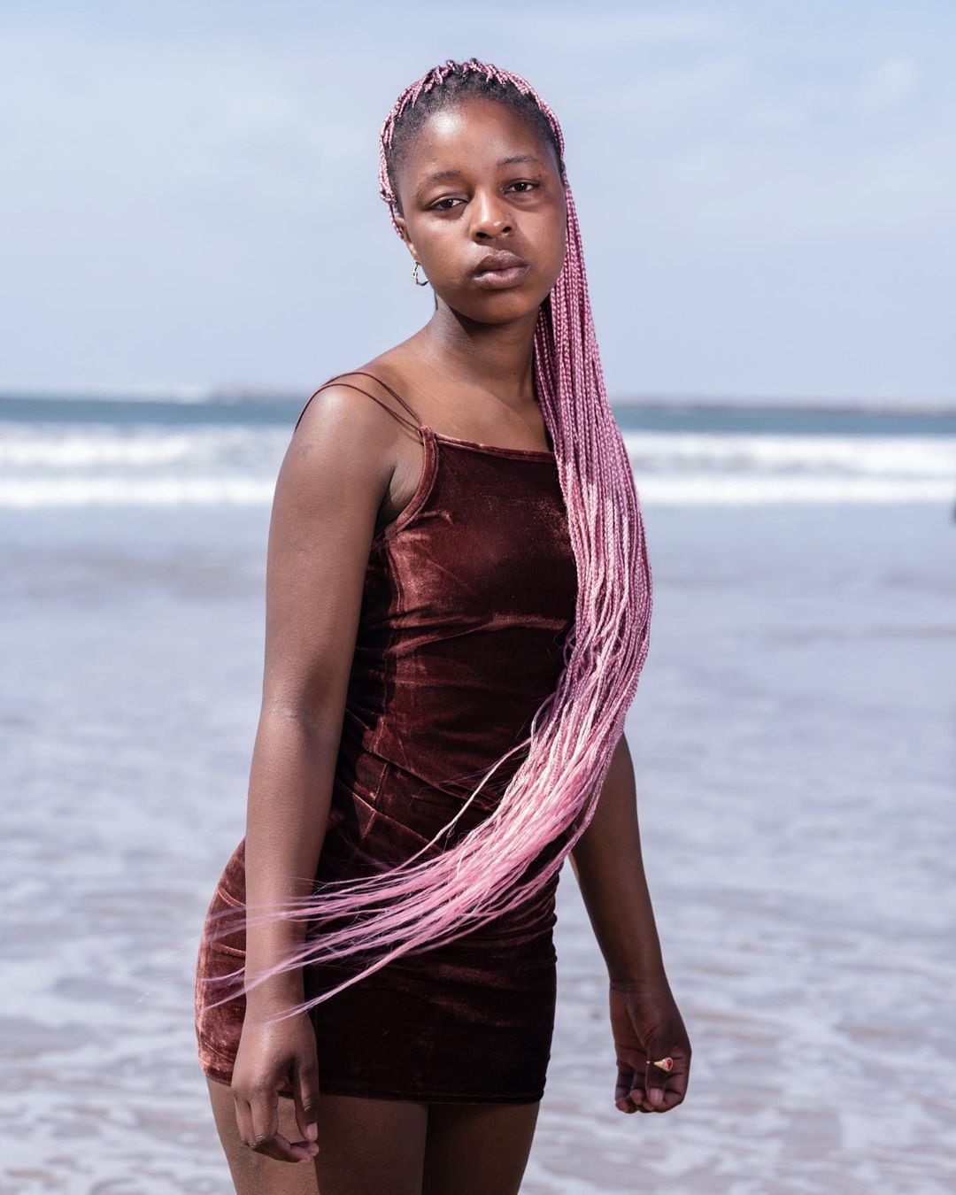 Young Black woman in a brown dress with pink braids standing on a beach with light blue ocean waves and a clear blue sky in daylight.
