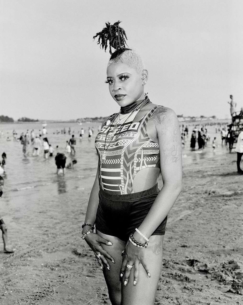 Woman in patterned swimwear and short shorts poses on sandy beach with crowd and water in background on bright day in black and white photo.