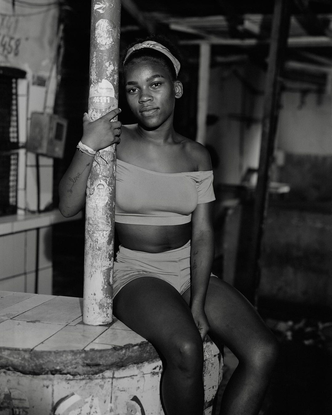 Monochrome portrait of a young woman in a two-piece outfit sitting and holding onto a pole indoors, looking directly at the camera.