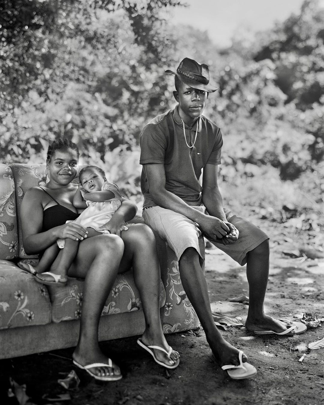 Monochrome portrait of a family of three outdoors: a woman holding a baby and a man in a cowboy hat sit outside on a patterned couch.