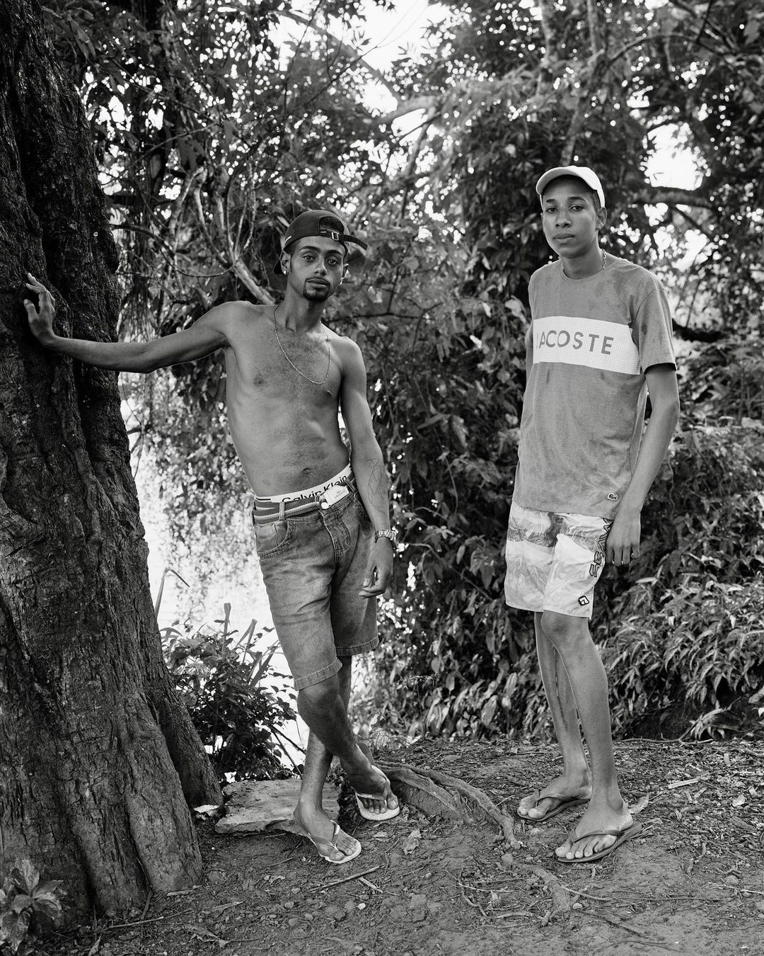 Two shirtless men stand next to a tree outdoors in a black and white shot. One is leaning against a tree, while the other is next to him.