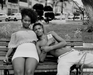 A black man and woman sitting together on a wooden park bench, captured in monochrome by Wayne Lawrence.