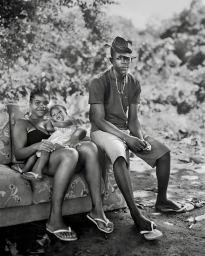 Black-and-white portrait of a family seated on an outdoor sofa with a man, a woman holding a baby, and dense foliage in the background.