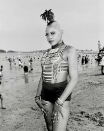 Black woman with styled hair standing on a sandy beach wearing a patterned swimsuit and dark shorts, photographed by Wayne Lawrence.