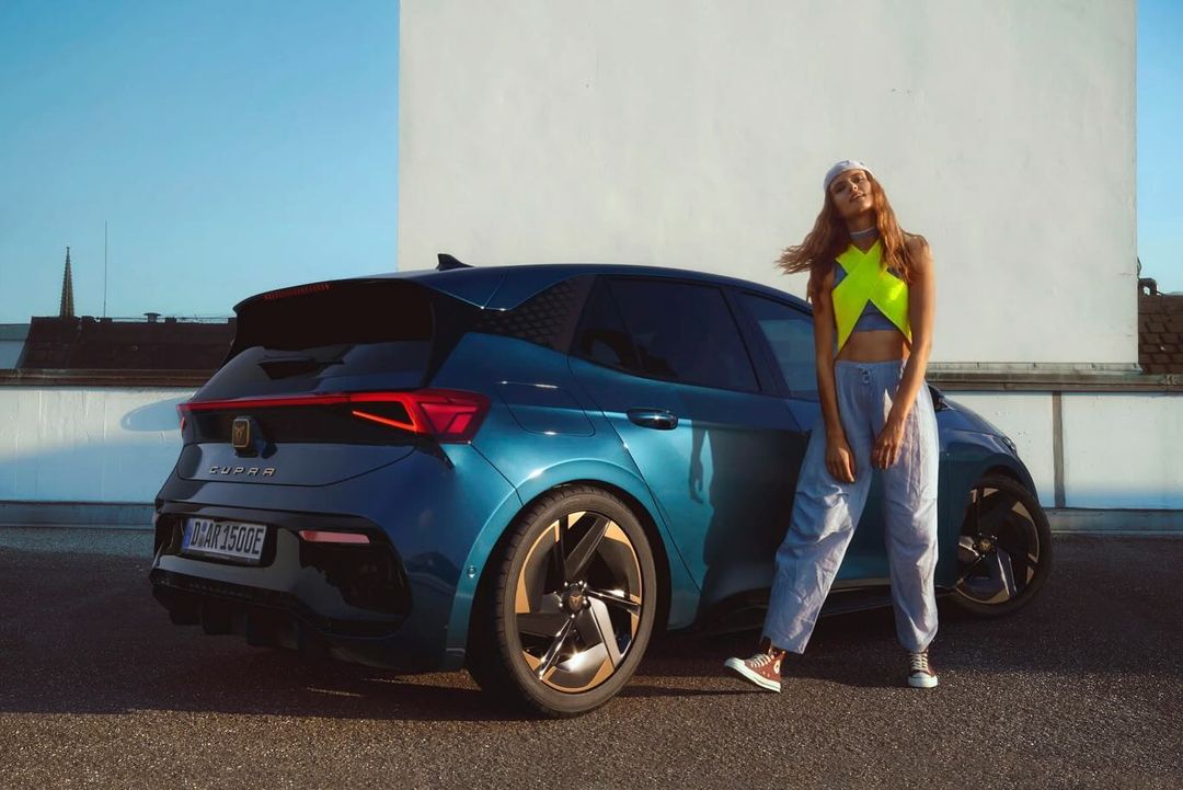 A teal Cupra electric car is parked outdoors, with a young adult female model standing next to it in a bright neon yellow top.