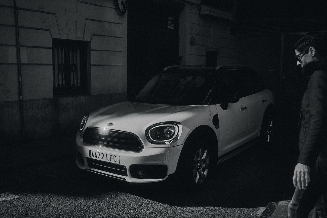 A white Mini Cooper Countryman parked on a city street at night, next to a pedestrian in a dark coat, in a black and white photograph.
