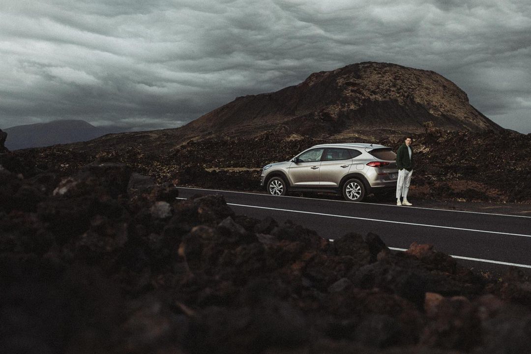 A silver SUV parked on an asphalt road, with a man leaning against it, set against a backdrop of rocky hills and cloudy skies.