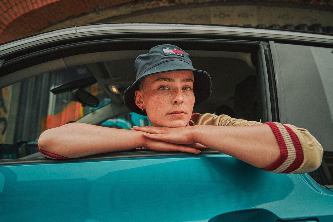 Close-up of a person looking out of a car window, wearing a navy bucket hat with a Tommy Jeans logo and tan jacket with striped sleeves.