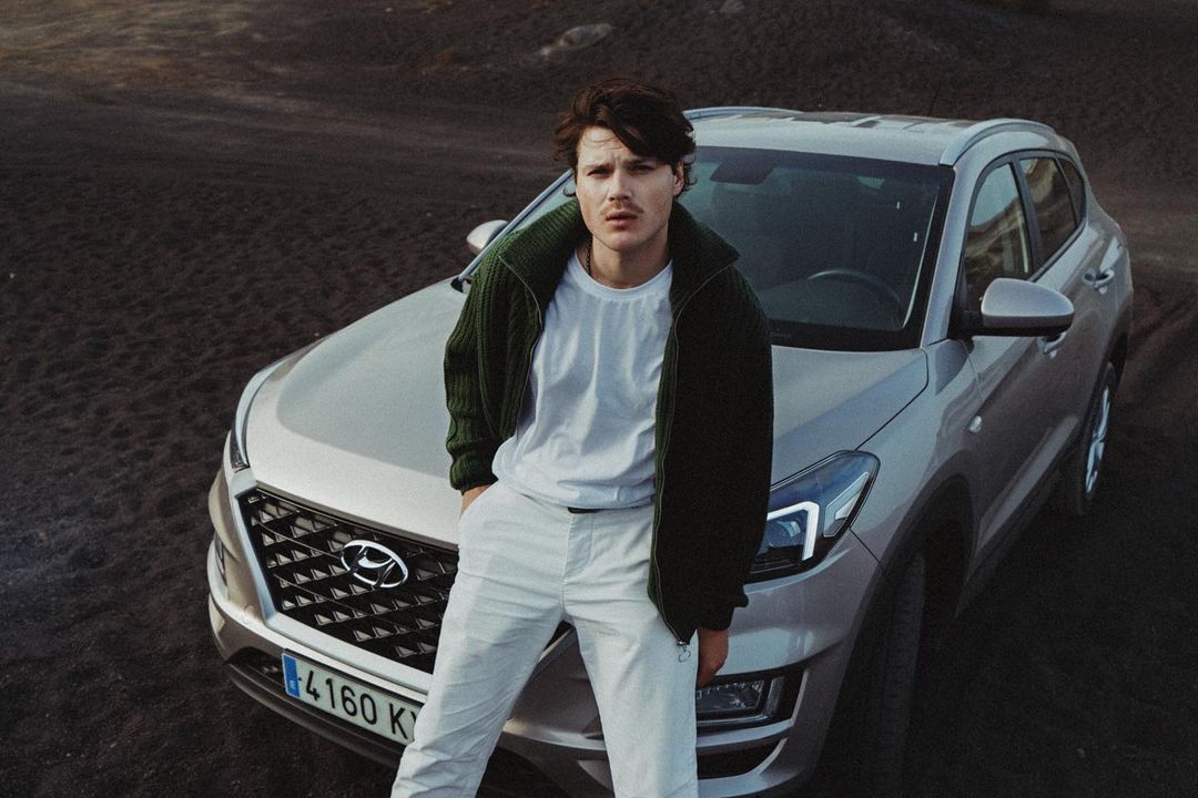Adult male poses with silver Hyundai Tucson SUV on black volcanic beach in olive green jacket and white pants, captured at eye-level.
