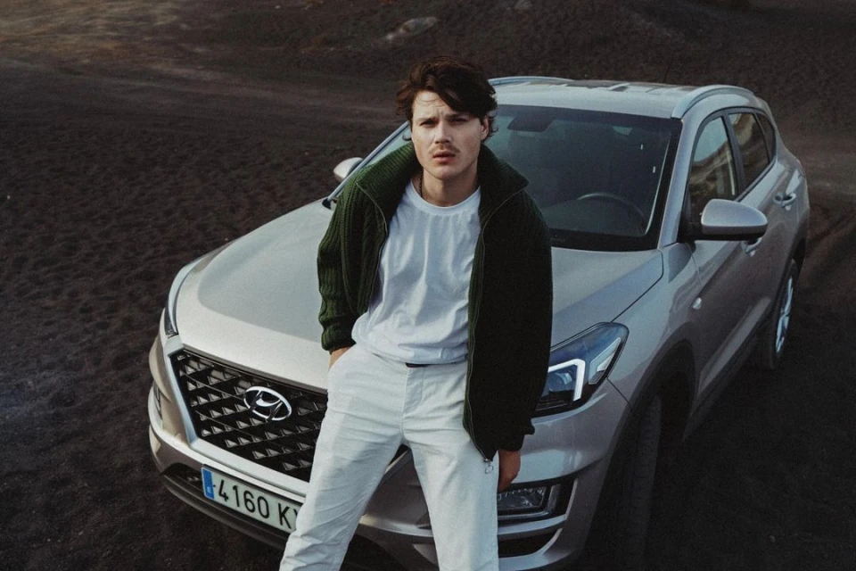 Adult male poses with silver Hyundai Tucson SUV on black volcanic beach in olive green jacket and white pants, captured at eye-level.