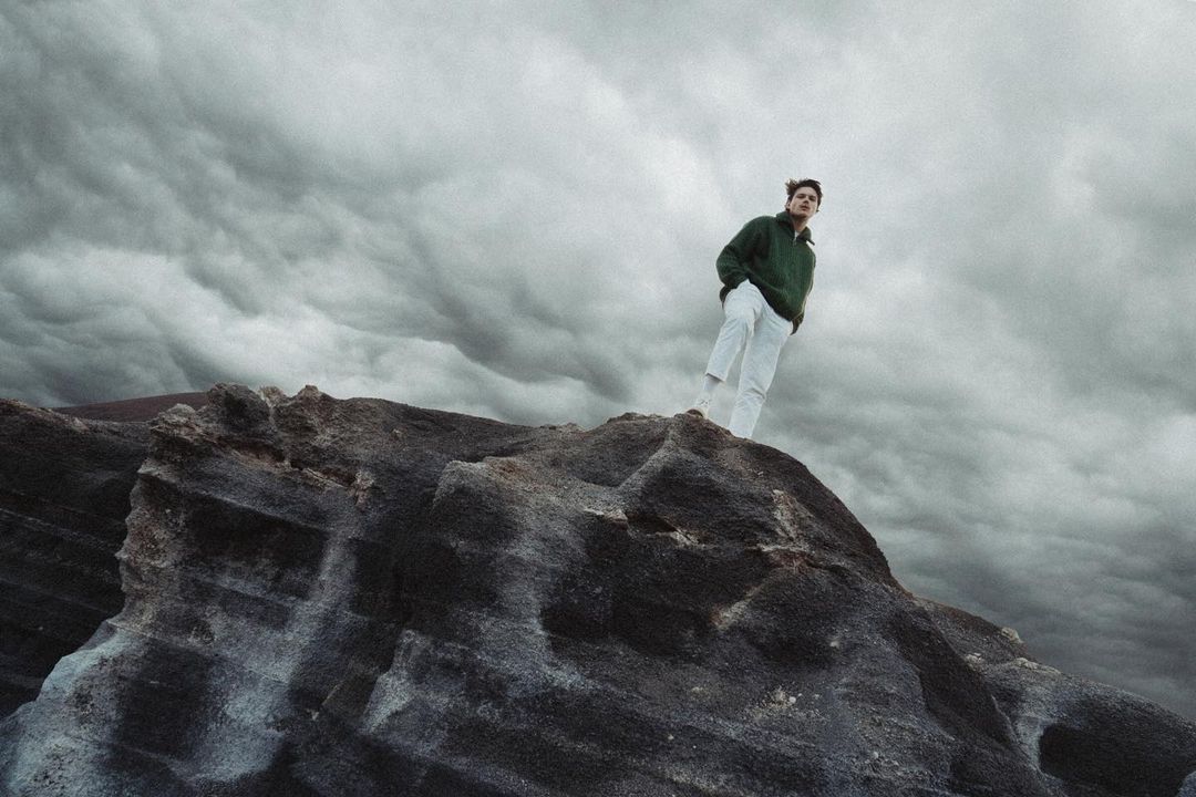 Adult stands on rocky peak beneath stormy sky, wearing a green jacket and white pants in a natural outdoor landscape.