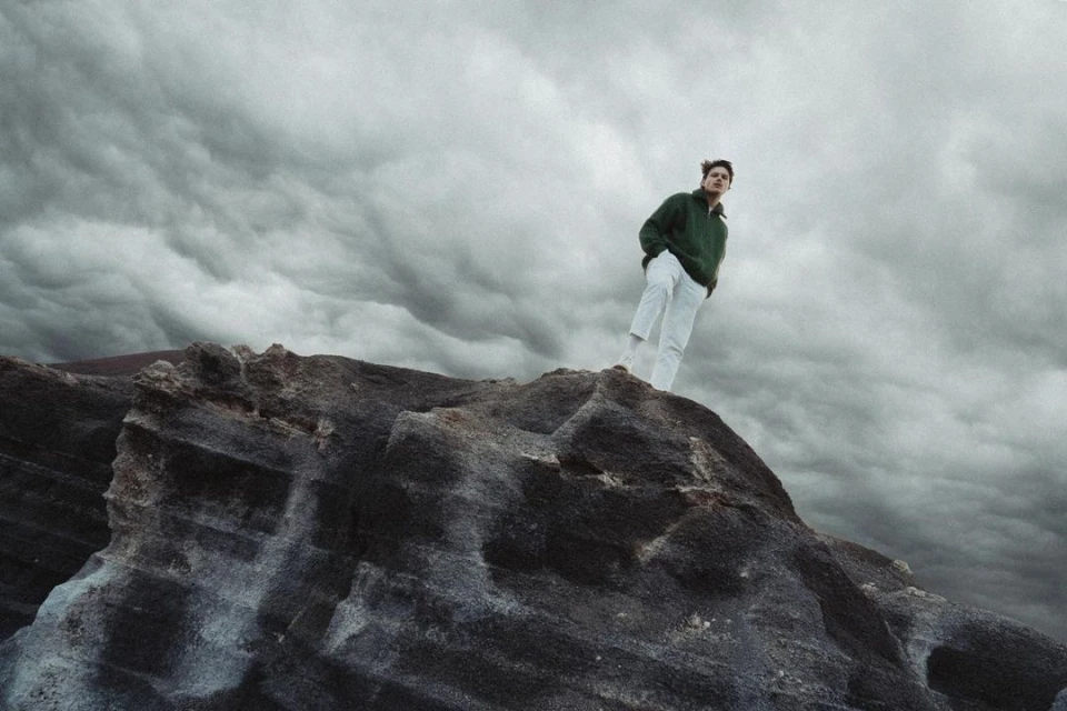 Adult stands on rocky peak beneath stormy sky, wearing a green jacket and white pants in a natural outdoor landscape.