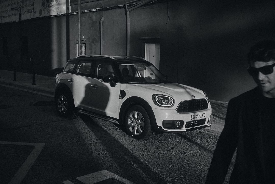 White Mini Cooper Countryman parked on a city street at night with a male figure partially visible in the foreground wearing sunglasses.
