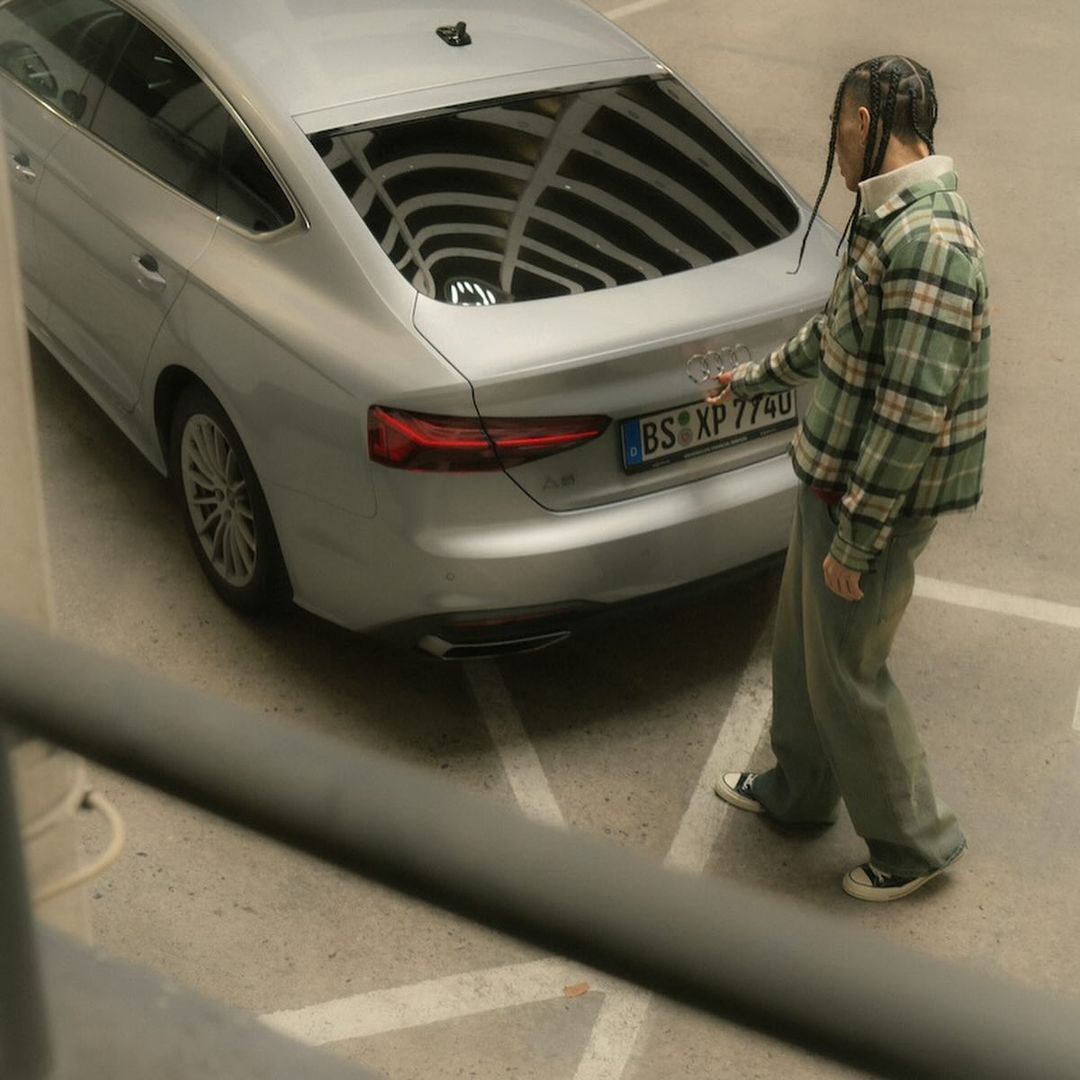 Silver Audi sedan parked with a person in a plaid shirt touching the car's logo in an urban parking garage setting.