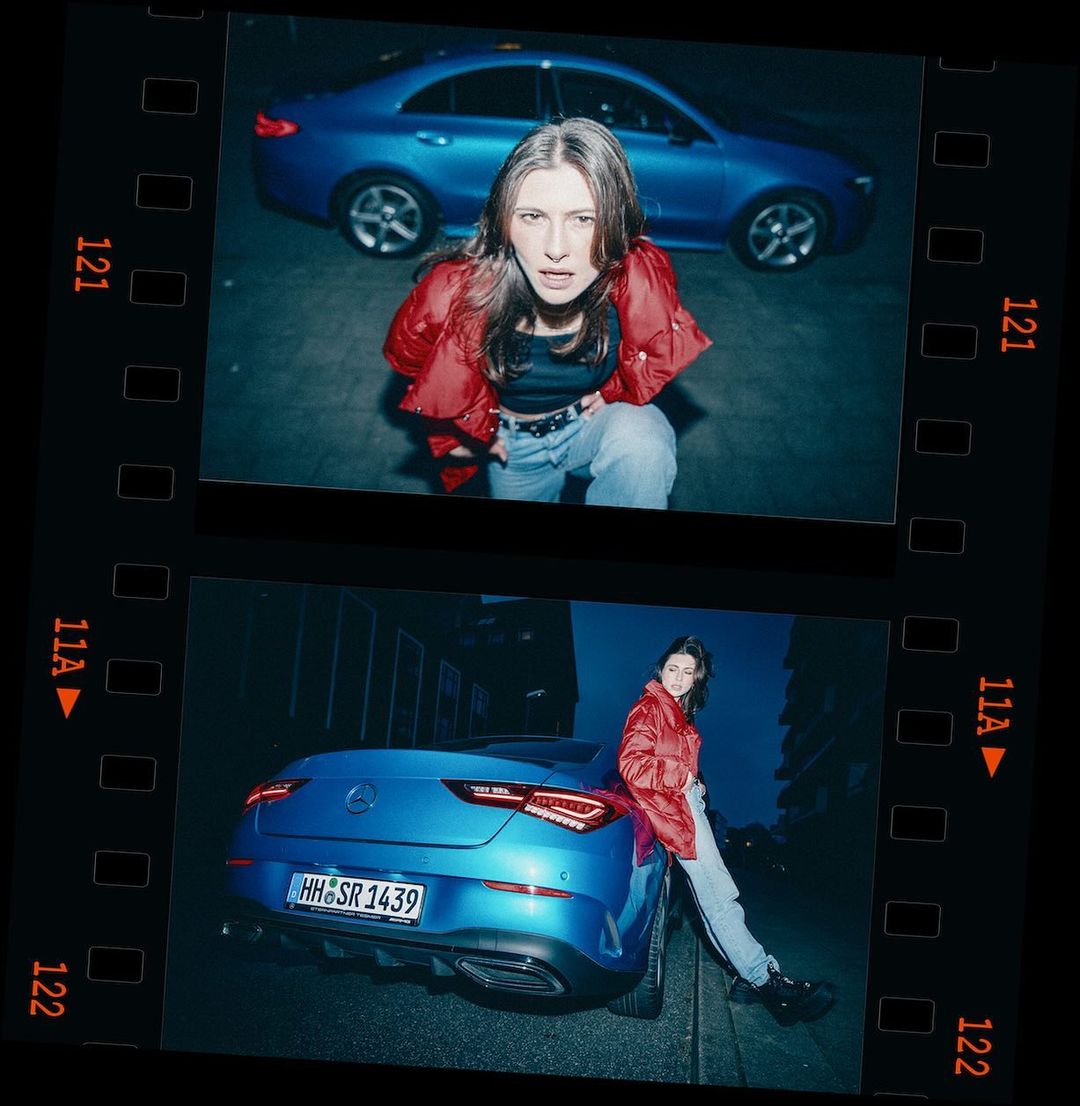 Young woman in a red jacket poses next to a blue Mercedes-Benz car with a European license plate in an urban setting at night.