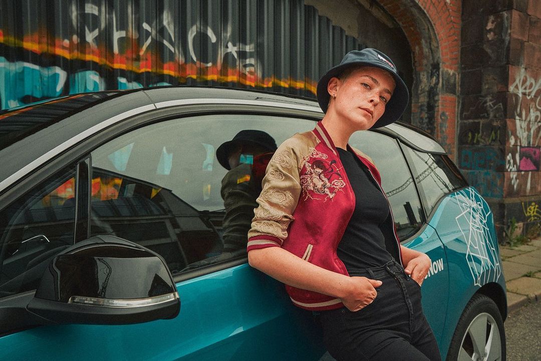 A young adult woman wearing a bucket hat leans against a teal electric car in front of a graffiti-covered brick wall in an urban setting.