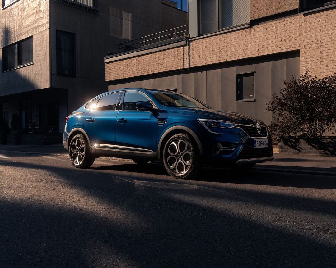 A blue Renault Arkana coupe SUV parked on a city street with a brick building in the background and sunlight casting shadows.