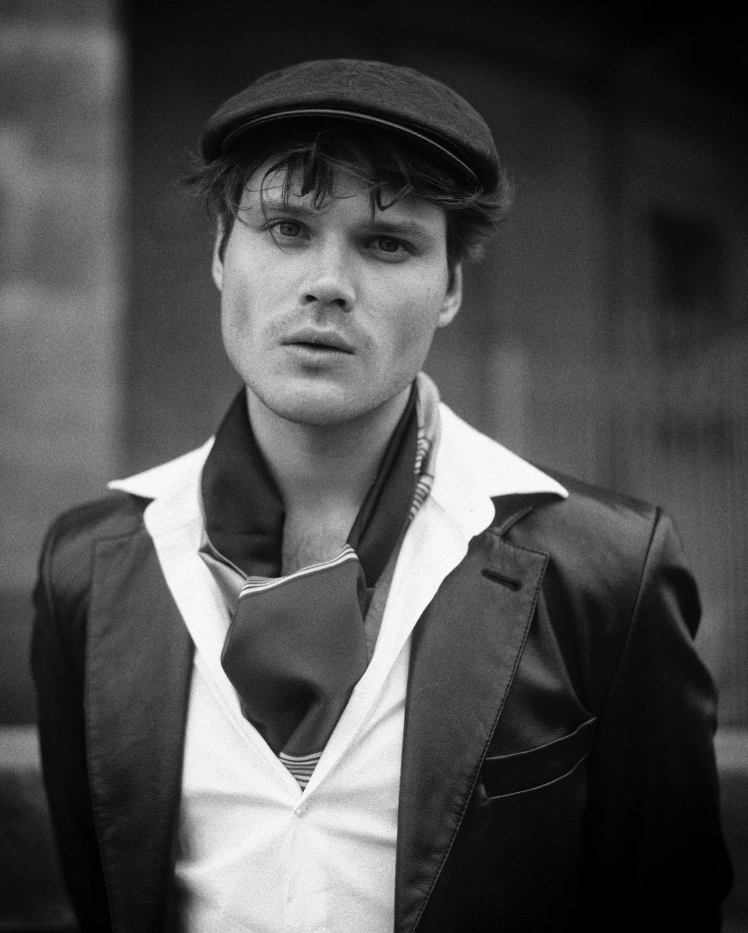 Monochrome portrait of adult male wearing a flat cap, blazer, and scarf, gazing at the camera against a blurred, neutral background.
