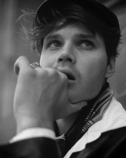 Black and white portrait of a contemplative young adult male wearing a cap, hand raised to his mouth, looking upward in thought.