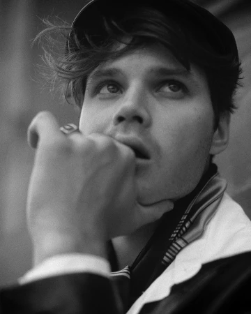 Black and white portrait of a contemplative young adult male wearing a cap, hand raised to his mouth, looking upward in thought.
