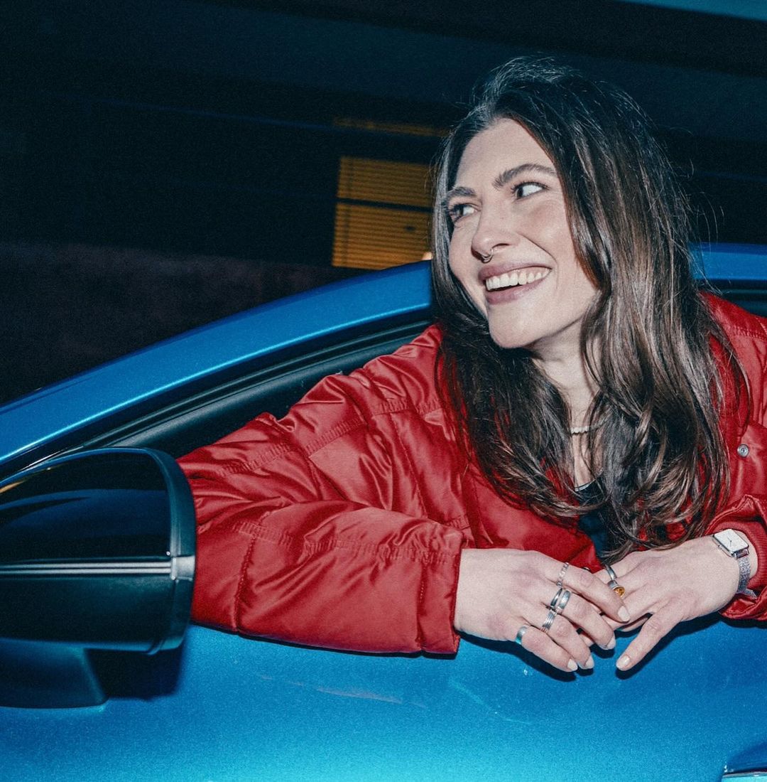 Brunette woman smiling while leaning out of the driver's seat of a blue car at night. She wears a red puffer jacket and rings.