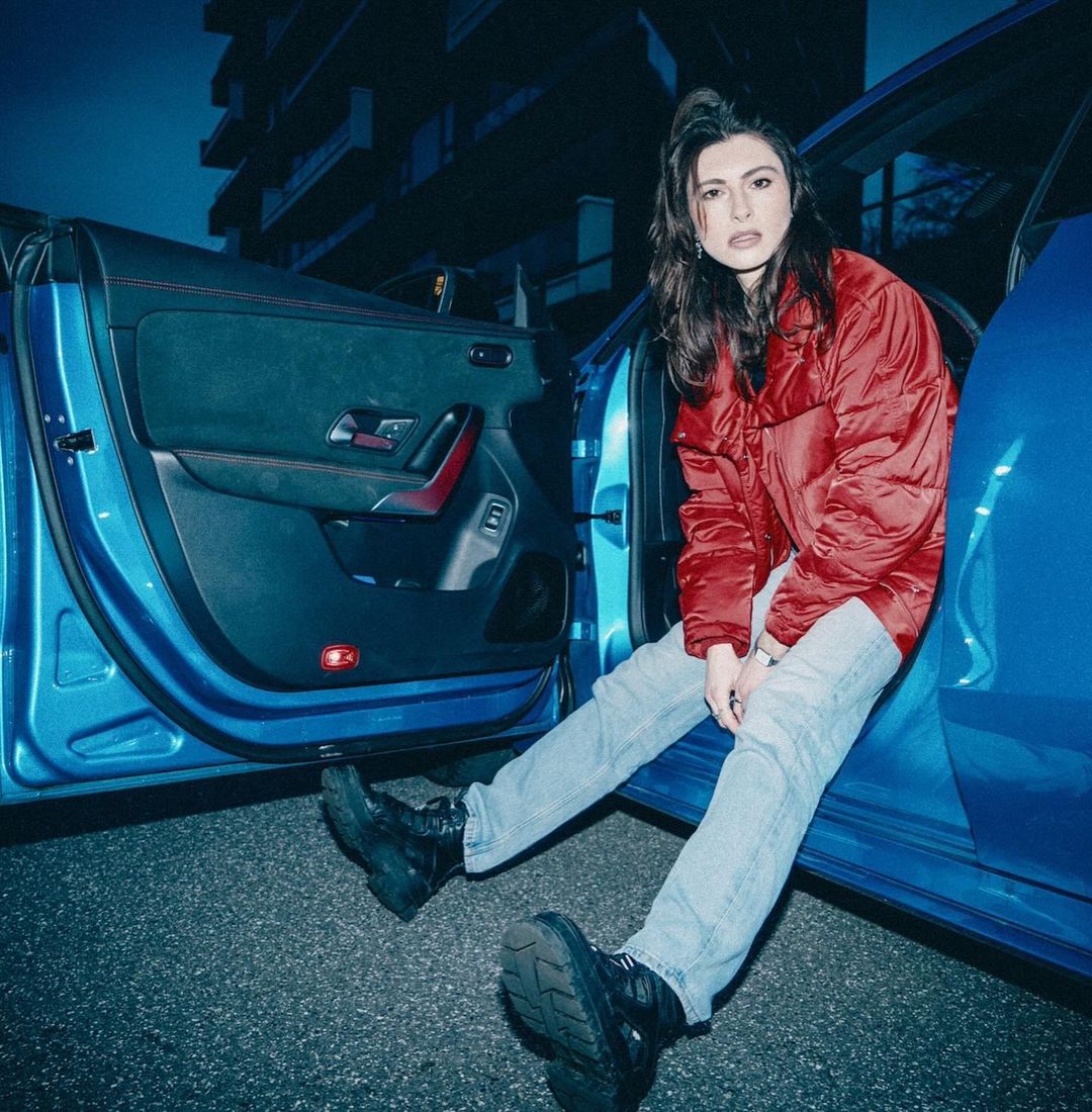 Young woman in a red puffer jacket sitting in the open doorway of a blue car in an urban setting at dusk or night.