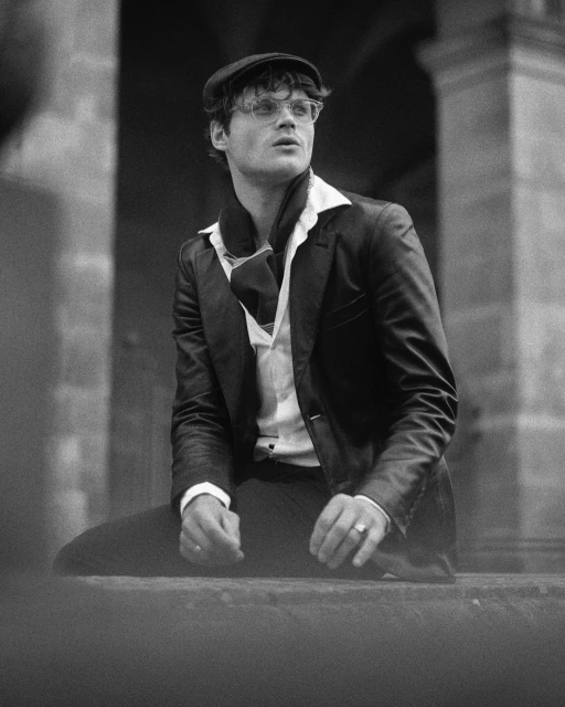 Monochrome portrait of a fair-skinned male model in a dark suit, newsboy cap, and glasses, looking up and to the right in an outdoor setting.