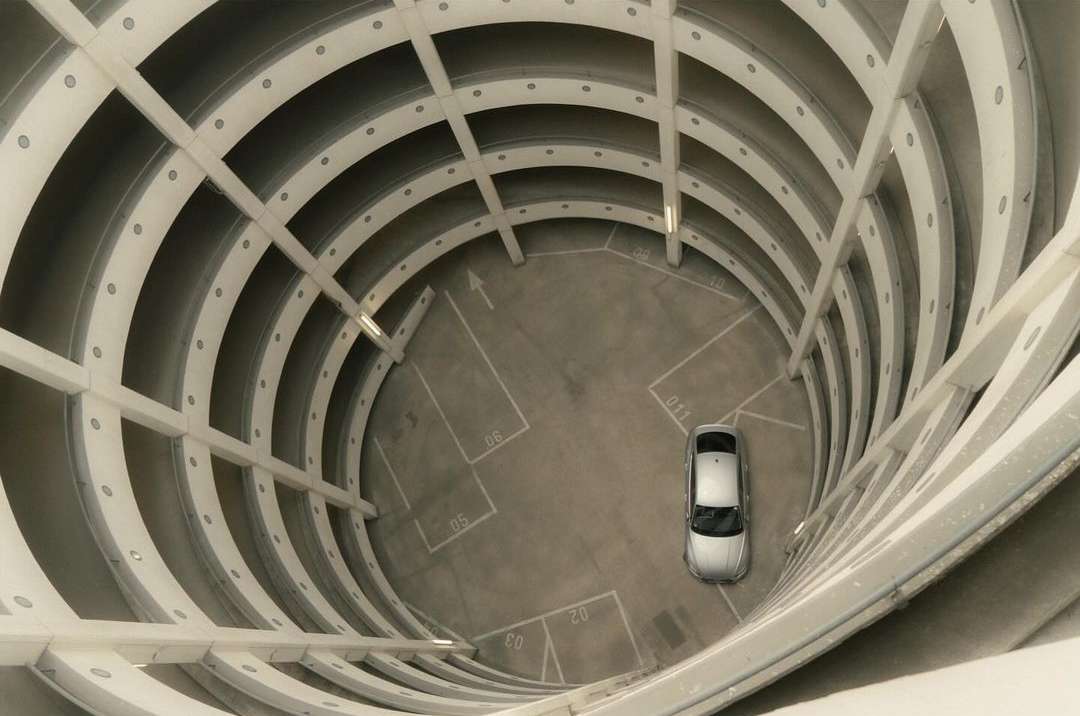 Elevated shot of a silver car parked inside a circular multi-story parking structure with concrete walls and clearly marked parking spaces.
