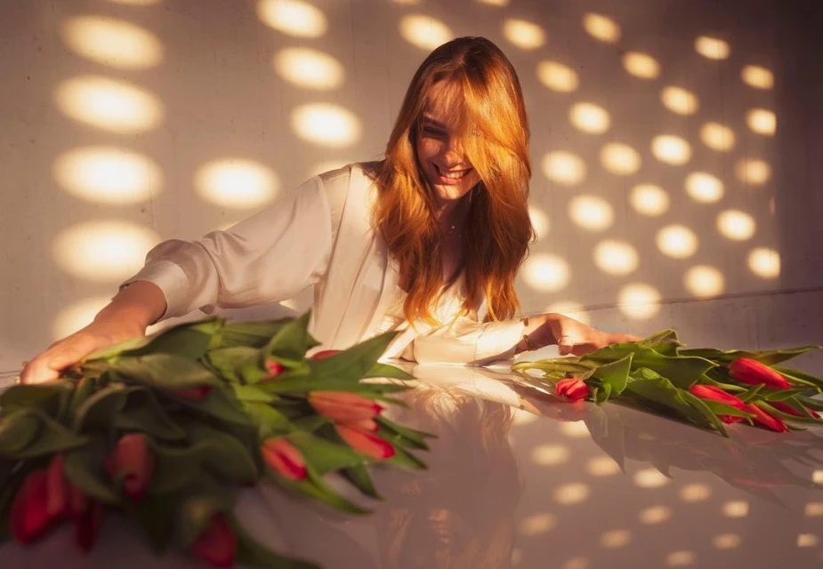 Woman with red hair in white jacket lying on table next to red tulips, bathed in warm, patterned light, against a textured background.
