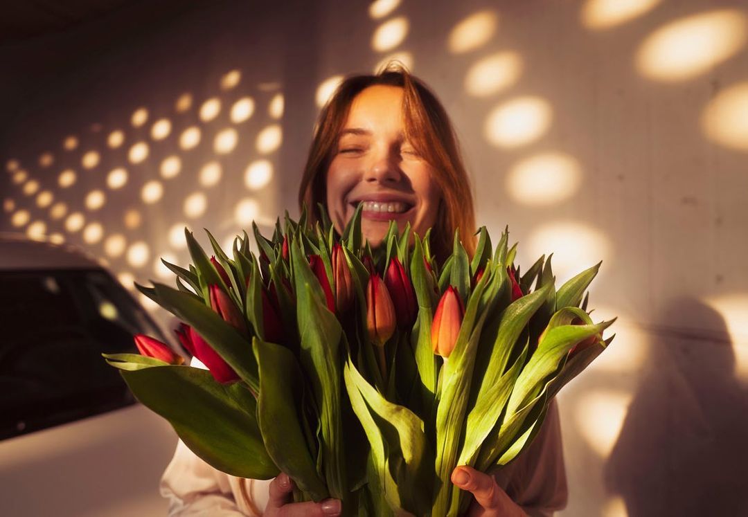 Smiling woman holds a large bouquet of red and orange tulips, illuminated by dappled sunlight and standing in front of a car.