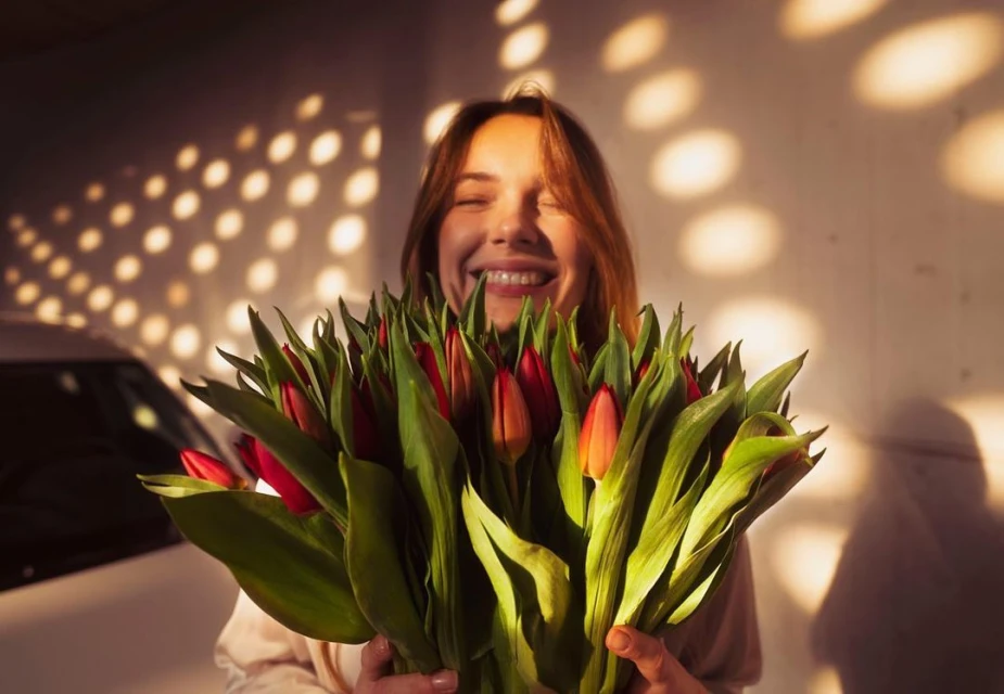 Smiling woman holds a large bouquet of red and orange tulips, illuminated by dappled sunlight and standing in front of a car.