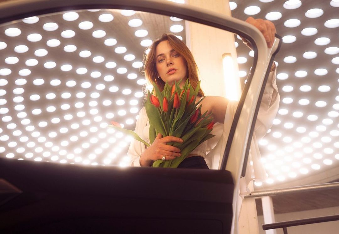 Elegant woman posing with red tulips beside a car in a white space with circular light pattern on the ceiling; fashion portraiture.