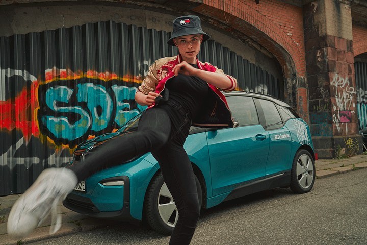 A young adult in a bucket hat kicks near a teal electric car under an overpass with graffiti in an urban setting with natural lighting.