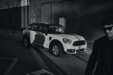Man wearing sunglasses walks past a parked white crossover car on a dark city street at night