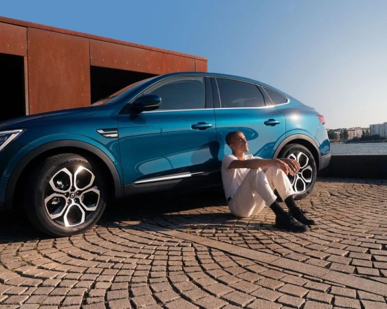 Man sitting on cobblestone ground leaning against a blue car with rusty metal panels in the background