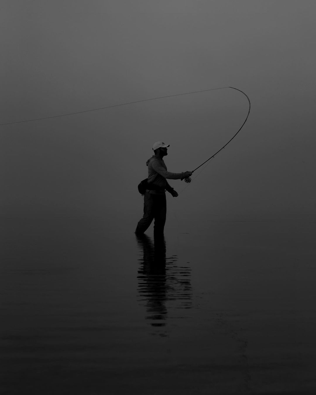 Monochrome photo of a man fly fishing while standing in water, casting his line with a curved rod under dark, overcast lighting.