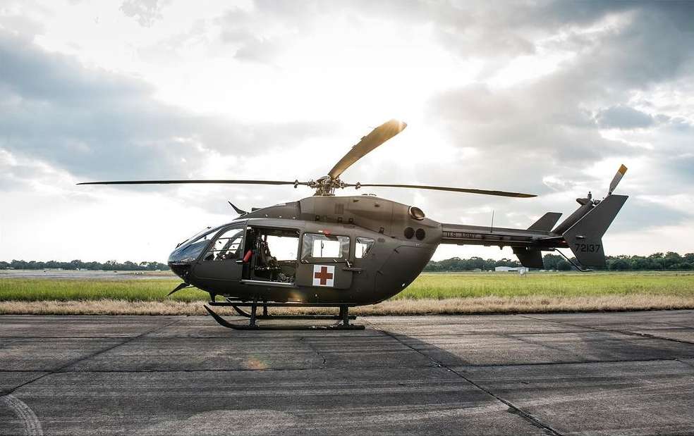 Olive drab medical helicopter parked on concrete pad against cloudy sky. Exterior view with open door and visible red cross symbol.