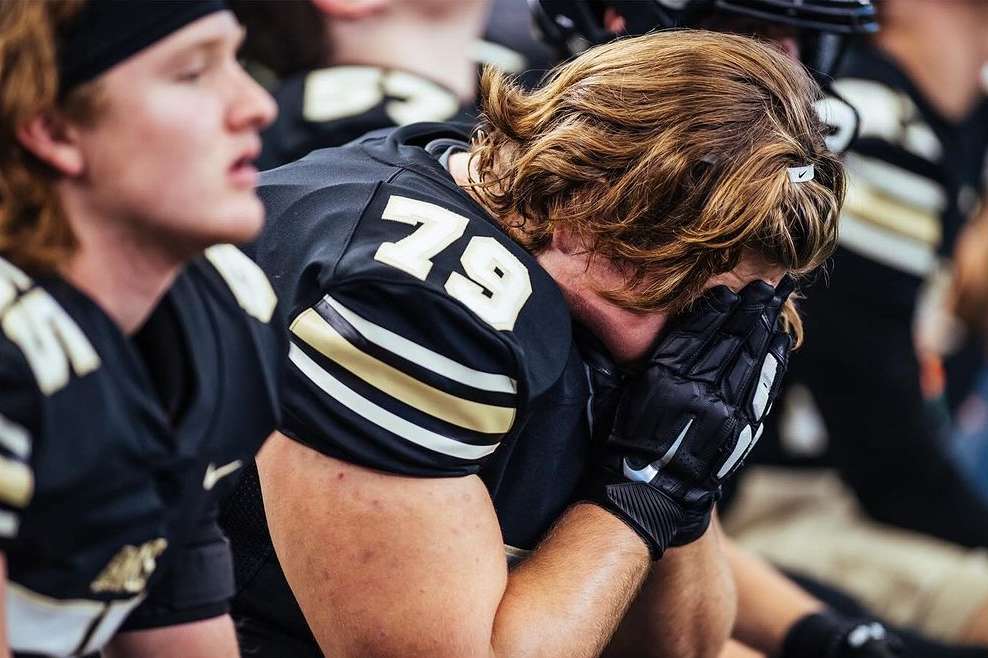 American football player in uniform sits on the bench, head in hands, showing dejection and disappointment after a loss.