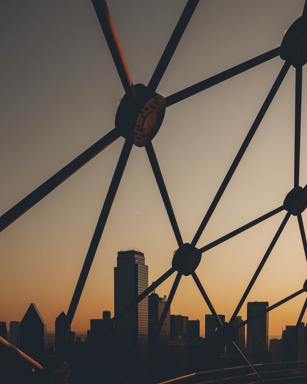 Abstract architectural view of a city skyline with silhouetted buildings seen through a metallic geodesic structure during golden hour.