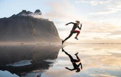 Woman running across a shallow reflective wetland with a dramatic dark mountain rising in the background at sunset.