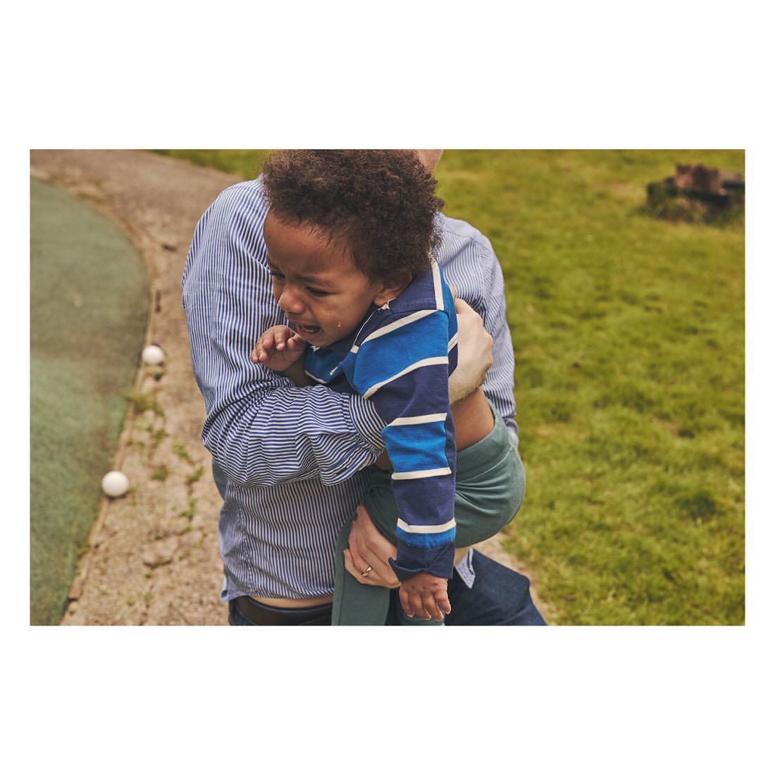 Toddler in striped shirt crying while held by an adult outdoors with a background of green grass and a white ball on a concrete path.
