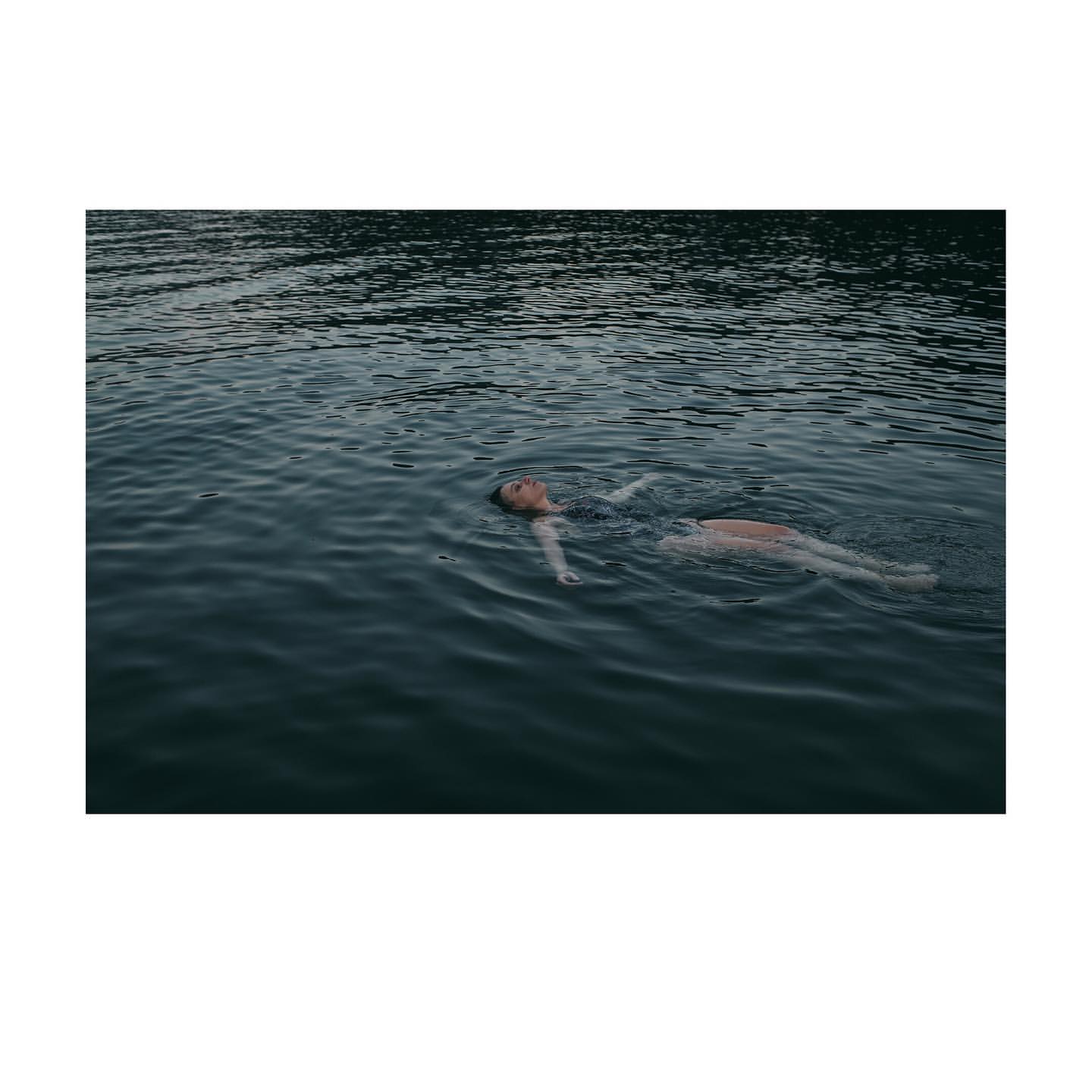Adult female floating in dark rippled water with forest background; eye-level, medium shot with dark color palette; summer season.