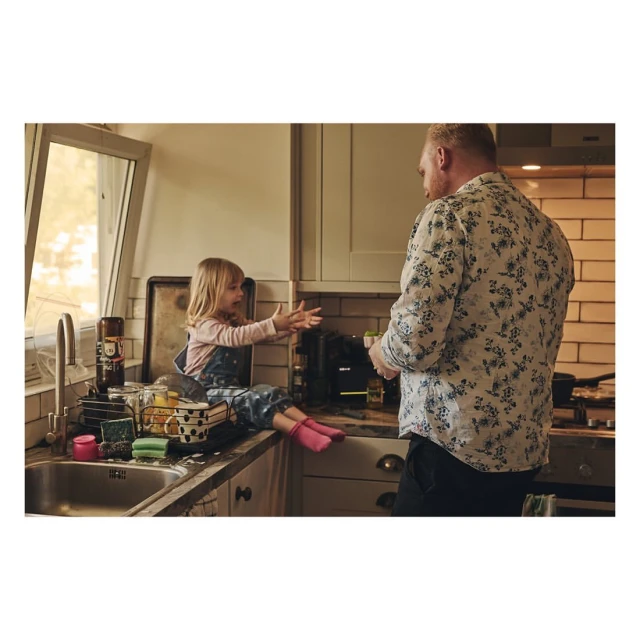 A blonde child sits on a kitchen counter near a sink, facing an adult in a floral shirt in a bright, homey indoor setting.