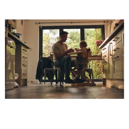 Father feeds his toddler in a wooden high chair in a warmly lit kitchen with large windows and dark cabinets. Happy family lifestyle.
