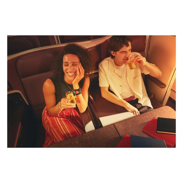 Two people in airplane seating enjoy drinks. An adult female smiles broadly as an adult male drinks from a glass in warm, inviting light.