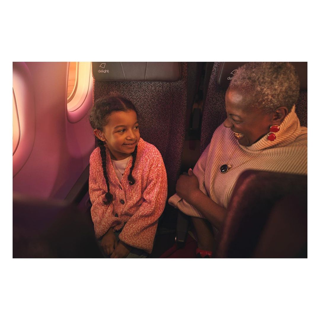 Smiling child and adult woman sitting on airplane, wearing pink and cream clothes, with window and interior in the background.