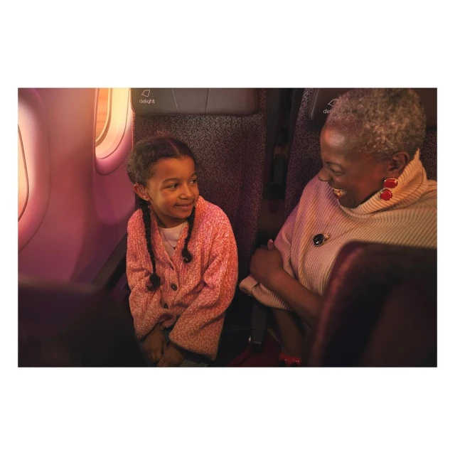 Smiling child and adult woman sitting on airplane, wearing pink and cream clothes, with window and interior in the background.