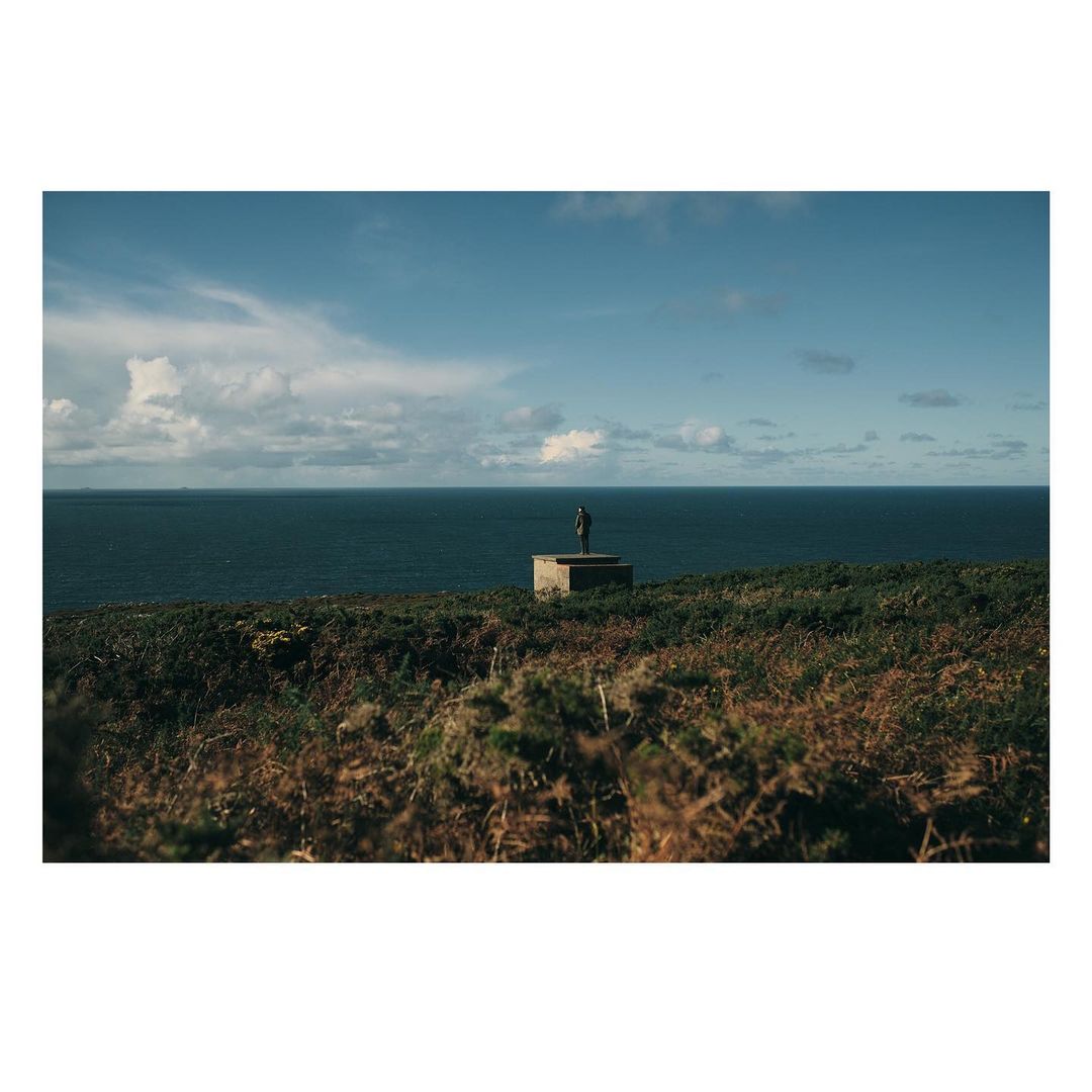 Outdoor shot features a distant sculpture on a stone base overlooking the sea, under a partly cloudy blue sky, with verdant foreground vegetation.