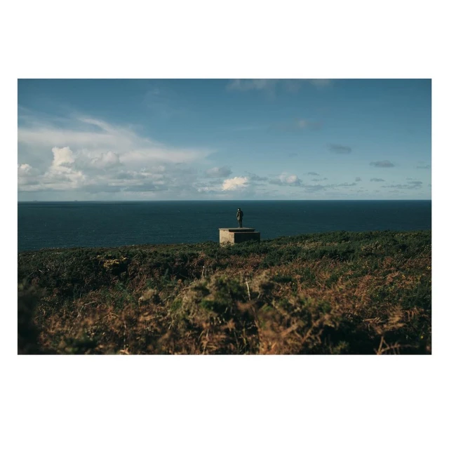 Outdoor shot features a distant sculpture on a stone base overlooking the sea, under a partly cloudy blue sky, with verdant foreground vegetation.